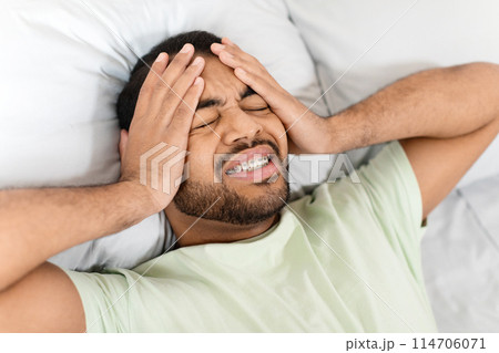 Sad sick african american millennial man laying in bed with closed eyes at home, touching head, tired black guy wake up with headache in the morning, suffering from chronic migraine, closeup 114706071