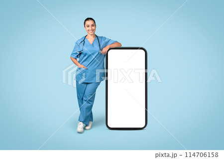 Confident woman nurse leaning on large, blank smartphone mockup for medical app demonstration, dressed in scrubs with stethoscope, on blue backdrop 114706158