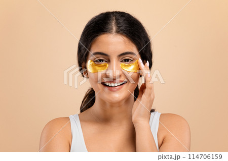 Happy beautiful young indian woman applying golden eye patches, cheerful eastern female enjoying moment of luxury skincare and self-care routine, looking at camera and smiling, beige background 114706159