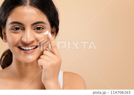 Cheerful young indian woman applying dab of moisturizing cream on her face, happy beautiful eastern female promoting skincare routine with radiant smile, standing against beige background, copy space Cheerful young indian woman applying dab of moisturizing cream on her face, happy beautiful eastern female promoting skincare routine with radiant smile, standing against beige background, copy space 114706163