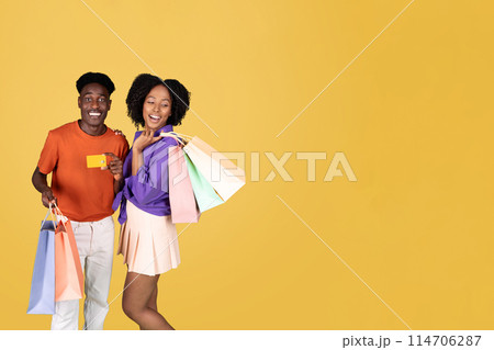 A young couple shopping, the woman in a purple blouse holding colorful bags and the man in an orange shirt showing a credit card, both smiling with satisfaction on a yellow background 114706287