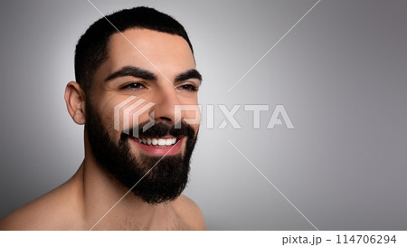 Closeup photo of positive handsome bearded middle eastern man posing on grey studio background, looking at copy space for advertisement and smiling, panorama. Men skin care concept. 114706294