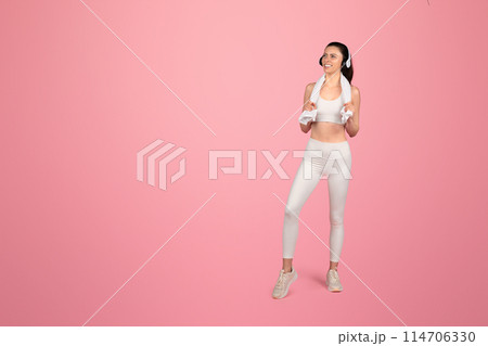 Smiling european fit woman in white sportswear with headphones around neck and towel over shoulders standing confidently on a pink background, radiating health and happiness Smiling european fit woman in white sportswear with headphones around neck and towel over shoulders standing confidently on a pink background, radiating health and happiness 114706330