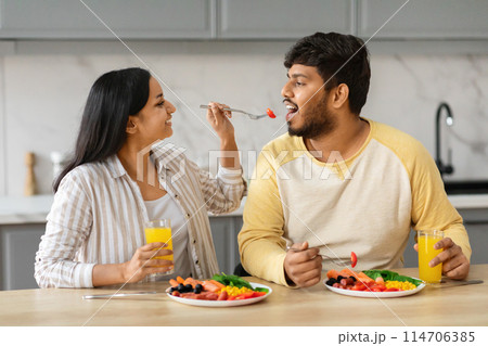 Pretty long-haired young indian woman feeding her husband, giving him fresh organic vegetables, modern kitchen interior. Millennial eastern couple spouses have breakfast together at home 114706385