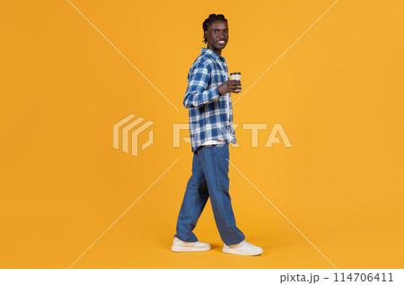 Relaxed young black man walking with takeaway coffee cup in hand, handsome african american guy dressed in plaid shirt and denim jeans posing on bright yellow background, copy space Relaxed young black man walking with takeaway coffee cup in hand, handsome african american guy dressed in plaid shirt and denim jeans posing on bright yellow background, copy space 114706411