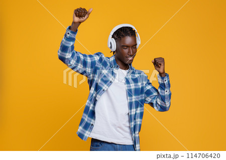 Positive young black man enjoying listening music, dancing and having fun, cheerful millennial african american guy wearing wireless headphones posing on yellow studio background, copy space 114706420