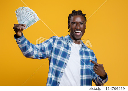 Happy Black Man Holding And Pointing At Dollar Cash Fan In His Hand, Cheerful Male Showing Money Banknotes While Standing Over Yellow Studio Background, Celebrating Financial Success And Profit 114706439