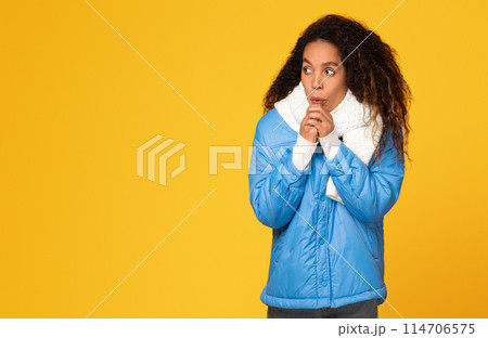 Young black lady in winter jacket shivers, hands clasped, as she looks towards an empty space on yellow background, suggestive of chilly weather 114706575