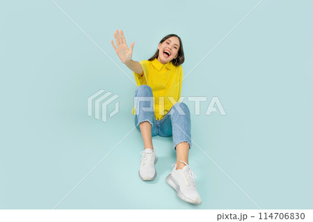 Joyful young brunette woman wearing stylish casual outfit sitting on floor and laughing, waving at camera, isolated on blue studio background, copy space 114706830