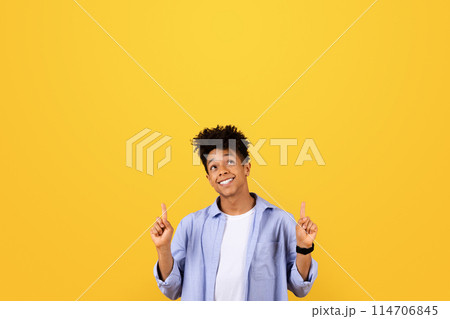 Cheerful black man in studio, stylishly dressed, smiling and pointing upwards at free space with both hands on vibrant yellow background, full of positivity Cheerful black man in studio, stylishly dressed, smiling and pointing upwards at free space with both hands on vibrant yellow background, full of positivity 114706845