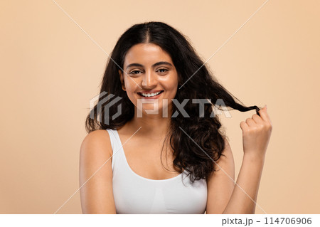 Happy young indian female showing her beautiful long hair after salon treatment and smiling at camera, attractive eastern female touching curl, standing isolated over beige studio background 114706906