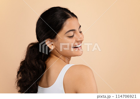 Beauty Concept. Smiling young Indian woman with luminous, unblemished skin standing gracefully against soft beige studio background, attractive eastern female enjoying her natural beauty, copy space 114706921