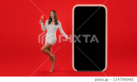 Young lady in festive attire presents blank smartphone screen template, holds a glass of sparkling wine in hand, symbolizing communication and celebration against red studio backdrop. Panorama Young lady in festive attire presents blank smartphone screen template, holds a glass of sparkling wine in hand, symbolizing communication and celebration against red studio backdrop. Panorama 114706948