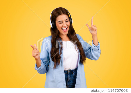 Glad teenager student lady use smartphone with headphones, sing song, enjoy music, lifestyle and fun, show peace sign, isolated on yellow background. Free time, relax app 114707008
