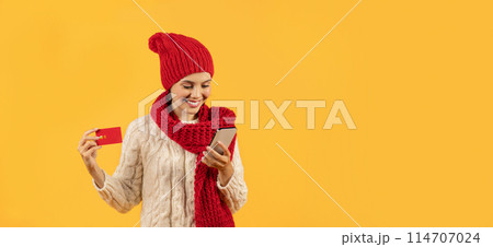 Cheerful young lady in winter scarf and hat using smartphone and credit card for online holiday shopping, paying in financial app, studio shot on yellow background. Free space for text. Panorama 114707024