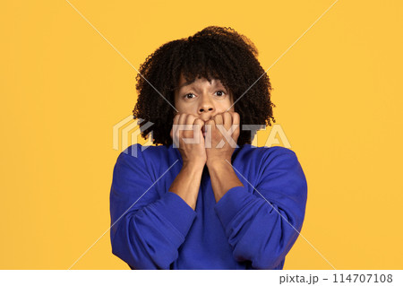 Anxious young black guy biting his nails and looking at camera, scared millennial african american man feeling worried, having panic attack, standing isolated on yellow studio background, copy space 114707108
