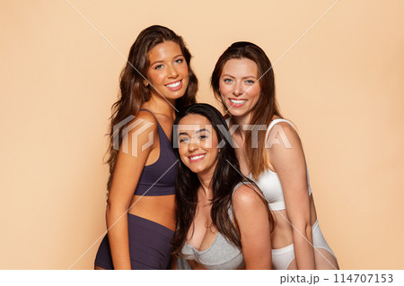 Three happy young international women in casual underwear together, smiling, embracing from behind, isolated on beige background. Friendship and togetherness, spa, beauty care Three happy young international women in casual underwear together, smiling, embracing from behind, isolated on beige background. Friendship and togetherness, spa, beauty care 114707153