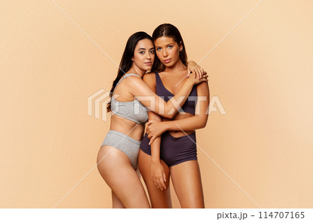 Two young european and latin women in sporty underwear pose closely, portraying trust and comfort in each other company, against soft peach backdrop. Friendship, trust, comfort and body care 114707165
