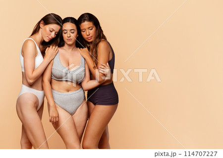Three diverse young women in comfortable undergarments share moment of closeness, offering solace and support on warm backdrop. Comfort, support, friendship and diversity 114707227
