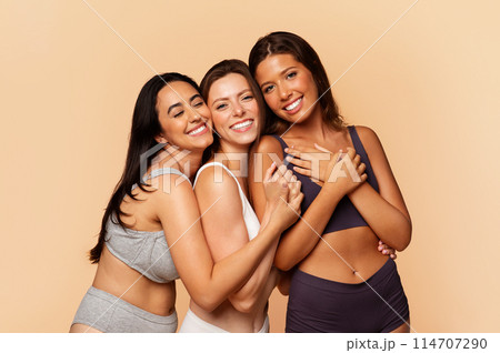 Three joyful international friends in casual underwear embracing and laughing, embodying friendship, diversity, isolated on light background. Lifestyle, body, beauty care 114707290