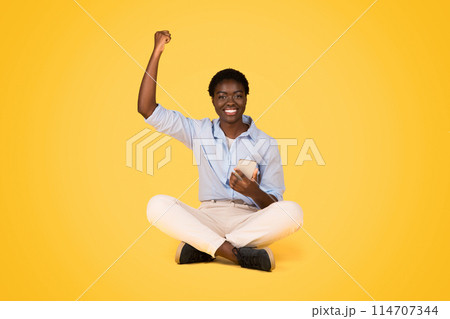 Cheerful young black woman student making win sign while using phone, isolated on yellow background, celebrate success, victory, sit on floor. Good work, game app, lottery 114707344