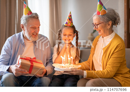 A young girl adorned with a birthday hat is sitting between her grandparents, who are smiling affectionately at her as they present a birthday cake with lit candles and a wrapped gift A young girl adorned with a birthday hat is sitting between her grandparents, who are smiling affectionately at her as they present a birthday cake with lit candles and a wrapped gift 114707696