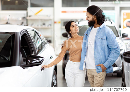 Indian couple standing next to a car in a parking lot. They are both looking towards the camera, with the car door open and the trunk slightly ajar. The man is holding car keys in his hand. Indian couple standing next to a car in a parking lot. They are both looking towards the camera, with the car door open and the trunk slightly ajar. The man is holding car keys in his hand. 114707760