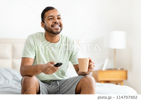 A cheerful African American man in casual clothing, sits comfortably on his bed holding a coffee mug in one hand and a remote control in the other. A cheerful African American man in casual clothing, sits comfortably on his bed holding a coffee mug in one hand and a remote control in the other. 114707802
