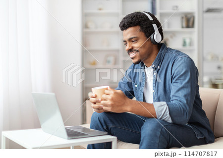 A cheerful young African American man is sitting comfortably on a white couch, donning a pair of headphones and handling a cup in his hands, enjoying music or engaging in a virtual conversation A cheerful young African American man is sitting comfortably on a white couch, donning a pair of headphones and handling a cup in his hands, enjoying music or engaging in a virtual conversation 114707817