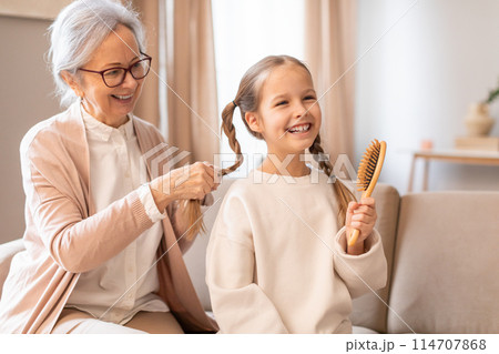 An older woman is gently combing the hair of a young girl, who is sitting still and looking content. Granny hands carefully detangle and style the girls hair, creating a tender moment between the two. 114707868