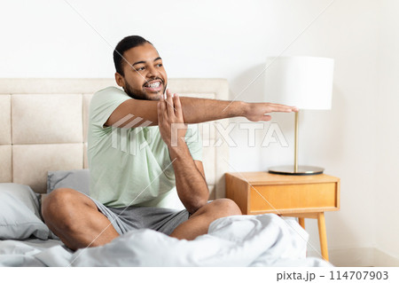 A young African American man is sitting on a bed, wearing casual sleepwear and stretching with a content smile 114707903