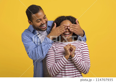 A smiling young African American man is standing behind a woman, playfully covering her eyes with his hands as she reacts with a joyful expression of surprise and anticipation 114707935