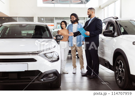 A couple of individuals are standing in front of a row of parked cars. The people appear to be conversing or taking a break. The cars are neatly lined up in the background. 114707959