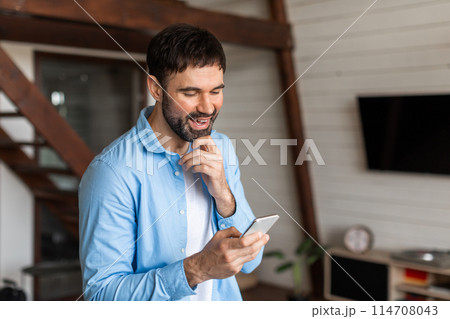 A cheerful young man with a beard is seen holding and looking at his smartphone with a smile, suggesting he has received good news A cheerful young man with a beard is seen holding and looking at his smartphone with a smile, suggesting he has received good news 114708043