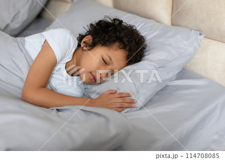 A small African American child peacefully sleeping on a bed while hugging a fluffy pillow. The child is nestled in blankets, and their eyes are closed in deep slumber. 114708085