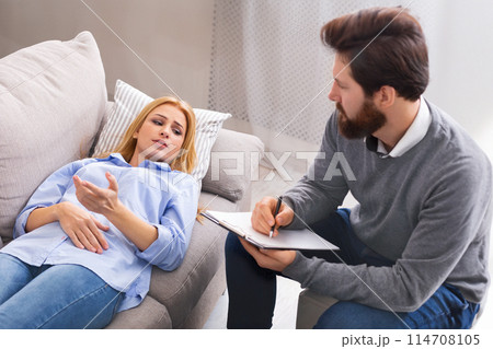 A young woman lies on a couch, looking pensive and speaking, while man therapist with a beard attentively listens and takes notes on a clipboard. A young woman lies on a couch, looking pensive and speaking, while man therapist with a beard attentively listens and takes notes on a clipboard. 114708105