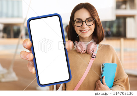 A woman student is holding up a modern smart phone with a blank screen in her hand. She appears to be looking at the screen, potentially preparing to use it for communication or information. 114708130