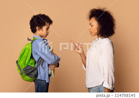 A young curly-haired African American boy carrying a green backpack attentively listens to a woman pointing her finger, possibly giving instructions or advice 114708145
