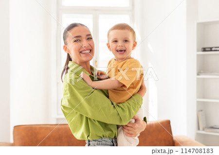 A woman is standing while holding a young child in her arms. The child appears to be looking up at the woman while she gazes down at them with a gentle expression. Both are dressed in casual clothing. 114708183