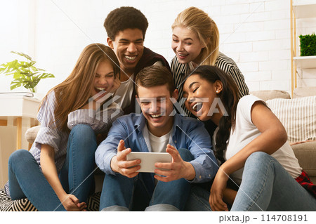 A cheerful group of multiethnic teenagers is clustered together on a couch, enjoying a moment of shared hilarity over content on a smartphone held by a smiling young guy A cheerful group of multiethnic teenagers is clustered together on a couch, enjoying a moment of shared hilarity over content on a smartphone held by a smiling young guy 114708191