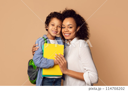 A cheerful African American mother lovingly hugs her young son, who is prepared for a day at school with his backpack and a bright yellow notebook A cheerful African American mother lovingly hugs her young son, who is prepared for a day at school with his backpack and a bright yellow notebook 114708202