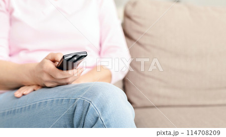 Cropped of woman is seated comfortably on a couch, holding a remote control in her hand. She appears relaxed and focused on the device, possibly changing channels or adjusting volume, copy space 114708209
