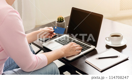 A woman sits at a cafe table engaging in online shopping or payment processing, while holding her credit card in one hand and typing on her laptop with the other. 114708247