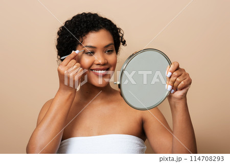 A young Hispanic woman is standing in front of a mirror, carefully applying makeup to her face. She is focused and precise, using various tools and products to enhance her features. 114708293