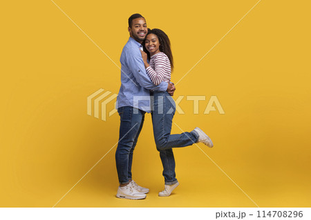 African American man and woman are standing close together, hugging each other tightly against a vibrant yellow background. Their arms are wrapped around each other as they share a warm embrace. 114708296