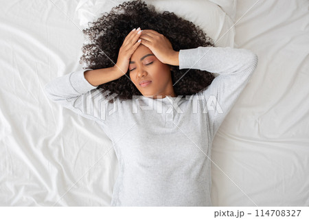 Hispanic woman is captured lying on her back in bed, with her hands placed on her forehead in a gesture that could indicate headache, stress, discomfort, or exhaustion Hispanic woman is captured lying on her back in bed, with her hands placed on her forehead in a gesture that could indicate headache, stress, discomfort, or exhaustion 114708327