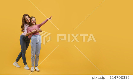 Mother and daughter dressed in casual wear, stand closely together against a vibrant yellow backdrop, pointing at copy space while they both smile brightly, sharing a lighthearted moment. Mother and daughter dressed in casual wear, stand closely together against a vibrant yellow backdrop, pointing at copy space while they both smile brightly, sharing a lighthearted moment. 114708353