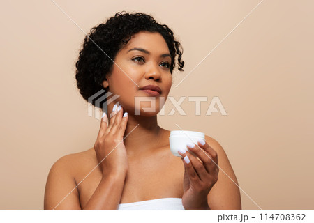 A young woman with curly hair is holding a jar of cream and gently applying it to her neck and jawline. She appears thoughtful and focused on her skincare routine A young woman with curly hair is holding a jar of cream and gently applying it to her neck and jawline. She appears thoughtful and focused on her skincare routine 114708362