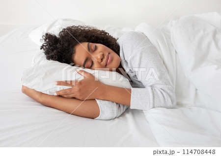 Hispanic woman is seen laying in bed, with a pillow under her head. She appears comfortable and relaxed, possibly resting or sleeping, side view 114708448