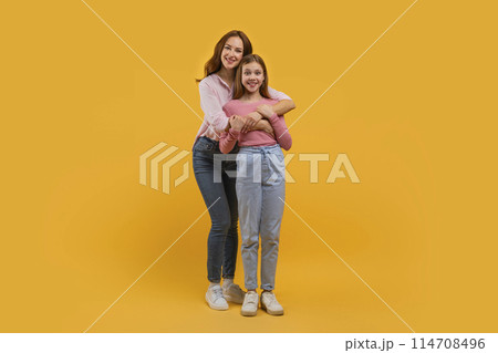 Mother and daughter stand closely with one embracing the other from behind. Both are smiling warmly and dressed casually in jeans and light tops against a vivid yellow backdrop. 114708496